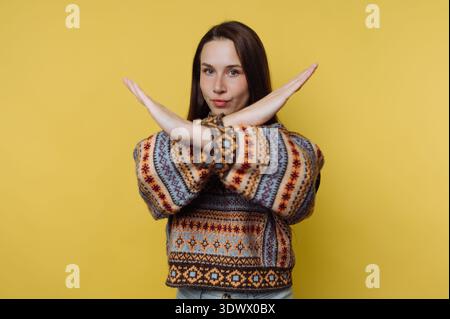 Une jeune femme se tient devant un mur jaune vif, affichant une position ludique mais affirmée avec les bras croisés. Elle porte un pull coloré th Banque D'Images
