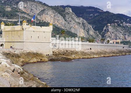Forteresse historique en pierre crème avec drapeau français le long de la côte rocheuse méditerranéenne et Mountains.Menton, France Banque D'Images