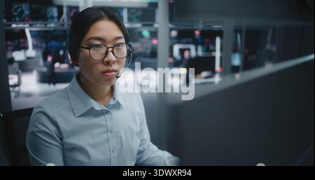 Jeune spécialiste asiatique de la chemise bleu vif, axé sur les tableaux de bord analytiques en temps réel. Femme portant un casque professionnel pour la communication instantanée. Experts de nouvelle génération en environnement d'investissement. Banque D'Images