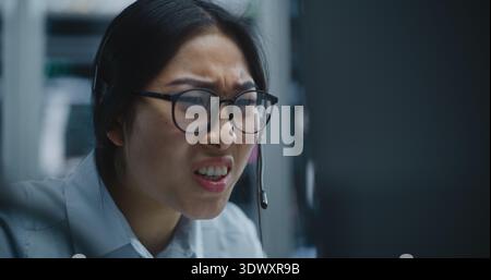 La négociante asiatique reçoit un appel via le casque tout en surveillant les données financières dans Modern Stock Office. Femme écoute Bad News à propos de la performance du marché, expression déplace l'inquiétude et la détresse. Banque D'Images