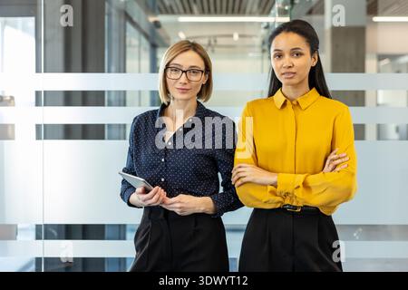 Deux femmes d'affaires confiantes debout côte à côte dans un bureau contemporain lumineux, représentant le partenariat, le travail d'équipe et la réussite professionnelle dans un environnement d'entreprise diversifié Banque D'Images