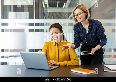 Deux femmes d’affaires souriantes collaborant et discutant du travail à l’aide d’un ordinateur portable et d’une tablette, favorisant le travail d’équipe et le développement professionnel dans un environnement de bureau contemporain Banque D'Images