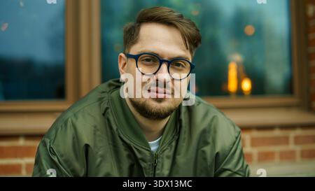 Portrait d'un beau jeune homme caucasien barbu dans des lunettes bleues et un blouson de bombardier vert, se relaxant à l'extérieur près d'un mur de briques. Banque D'Images