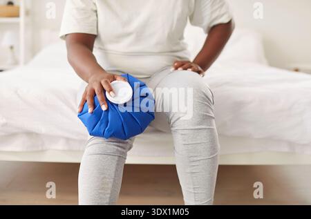 Femme afro-américaine assise sur le lit les mains touchant le genou avec le chauffage, se sentant blessée douleur articulaire Banque D'Images