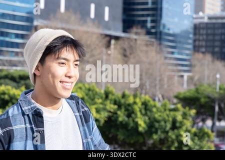 Adolescent asiatique portant un bonnet, une chemise à carreaux et un pull blanc, debout dans le parc par des bâtiments en verre Banque D'Images