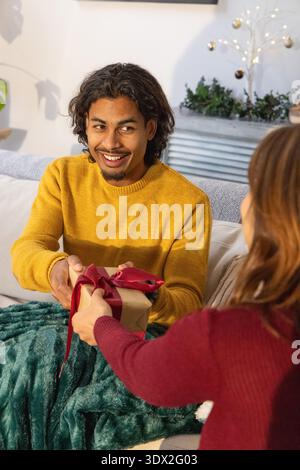 Couple échangeant cadeau, homme portant pull moutarde assis sur le canapé à la maison avec jeté vert Banque D'Images