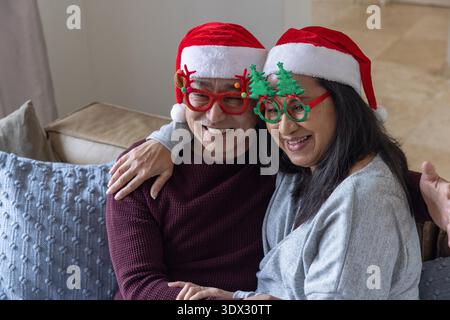 Couple senior diversifié souriant tout en portant des chapeaux de Père Noël et des lunettes de fête sur le canapé dans le salon. Festif, célébration, compagnie, chaleur, vacances, Joy, joyeuse || autorisation du modèle Banque D'Images