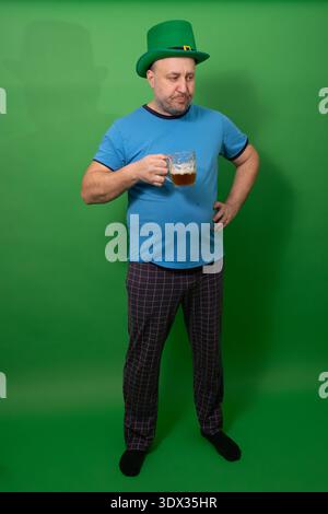 Homme dans le chapeau de leprechaun vert tenant le verre de pinte de bière mousseux avec la main sur la hanche. – Patricks Day pose humoristique et expression festive. Concept de célébration de bière de tradition de vacances irlandaise pour le plaisir de la fête. Banque D'Images