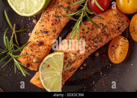 Steaks de saumon grillés cuits au four avec tomates, romarin, citron vert et épices gros plan sur assiette foncée. Carte parabolique vue de dessus. Banque D'Images