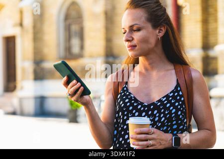 Jeune femme adulte avec du café pour aller en utilisant son smartphone dans une rue de la ville. Banque D'Images