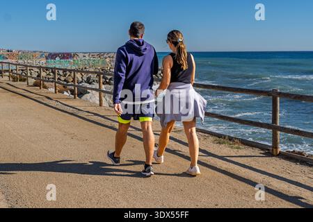 Un jeune homme et une jeune femme marchent ensemble le long d'un chemin de gravier au bord de l'océan par une journée ensoleillée. Banque D'Images