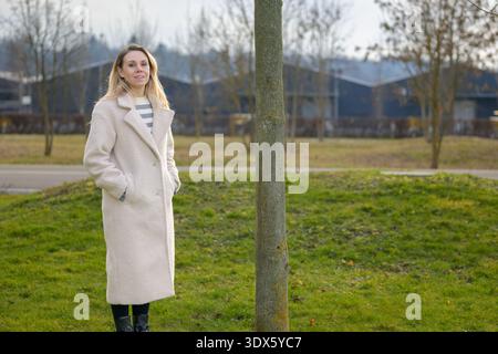 Femme blonde dans un manteau d'hiver léger debout dans un parc vert avec les mains dans les poches, l'air détendu et confiant, représentant le style de vie en plein air, lei Banque D'Images
