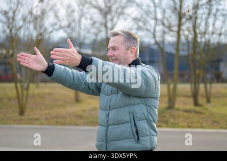 Homme mature souriant dans une veste verte étirant ses bras à l'extérieur dans un parc, profitant de l'air frais et de l'exercice léger par une journée ensoleillée. Banque D'Images