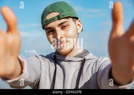 Un jeune homme à la barbe courte, portant une casquette verte et un sweat à capuche gris, étend ses bras vers la caméra, debout à l'extérieur contre un SK bleu vif Banque D'Images