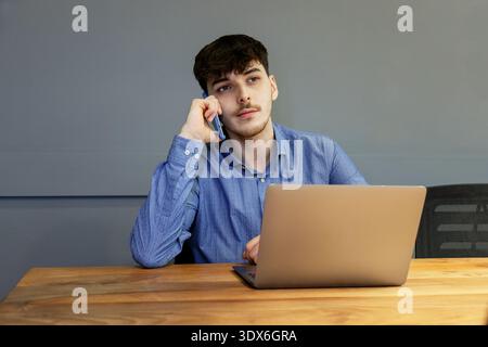Un jeune homme dans une chemise bleue est concentré alors qu'il travaille sur son ordinateur portable et parle au téléphone. Il semble s'occuper d'une affaire sérieuse dans un professeur Banque D'Images