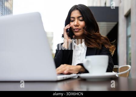 Une femme d'affaires hispanique travaille sur son ordinateur portable tout en parlant au téléphone dans un café en plein air. Elle est concentrée sur ses tâches, incarnant le professionnalisme et Banque D'Images