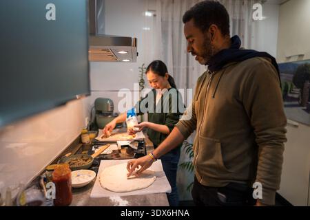 Un couple interracial profitant de la cuisine maison dans leur cuisine, préparant une délicieuse pizza maison ensemble. La femme ajoute des ingrédients, tandis que th Banque D'Images