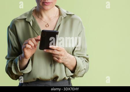 Une femme d'âge moyen pointe vers l'écran de son smartphone, portant une blouse vert clair et un pantalon gris sur un fond vert Uni. Calme et concentré, s Banque D'Images