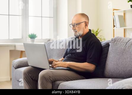 L'homme mûr ajuste les lunettes tout en travaillant sur l'ordinateur portable à l'intérieur du salon à la maison Banque D'Images