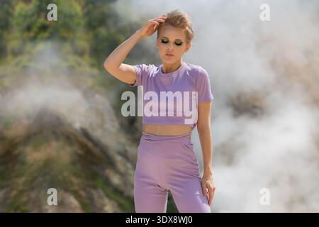 Une jeune femme à la mode avec une coupe pixie et un maquillage vert vibrant pose sereinement à l'extérieur. Portant une tenue lilas, elle se tient dans une lumière douce contre Banque D'Images