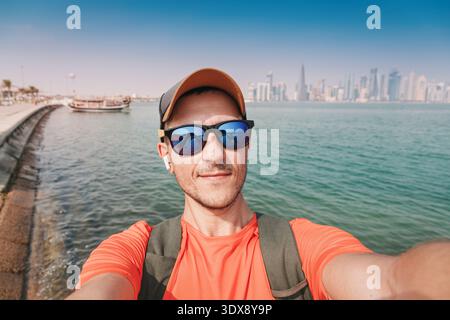 Homme souriant, portant des lunettes de soleil et un chapeau, prenant un selfie avec les gratte-ciel de West Bay de Doha et le front de mer de Corniche en plein jour Banque D'Images