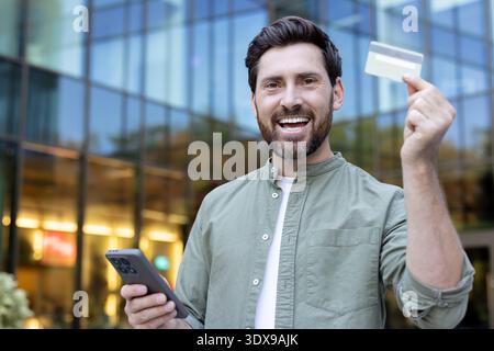 Homme heureux tenant une carte de crédit vierge et un smartphone, souriant tout en effectuant un paiement électronique ou un achat en ligne sur un fond de bâtiment flou moderne Banque D'Images