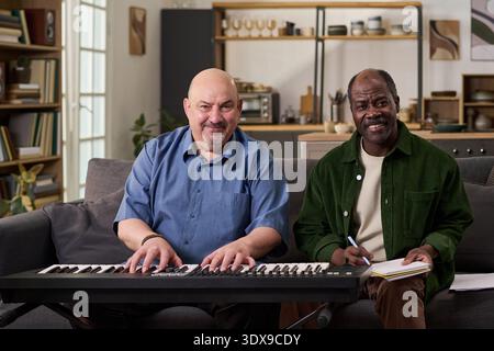 Homme caucasien d'âge moyen jouant du piano numérique assis à côté de l'homme noir d'âge moyen écrivant dans un cahier, à la fois souriant et regardant vers l'avant dans le cadre moderne du salon Banque D'Images