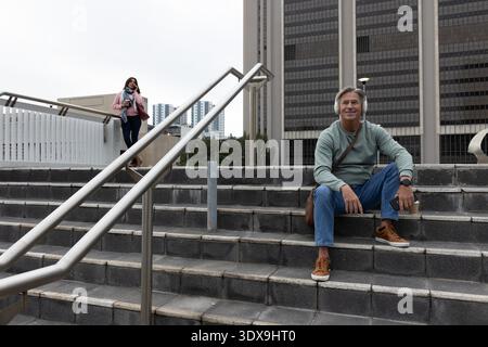 Homme senior assis sur les marches de la plaza portant des écouteurs et tenant le café pendant que la femme asiatique appelle. Piéton, urbain, navetteurs, escaliers, smartphones, écouteurs, sweat-shirt || autorisation du modèle Banque D'Images