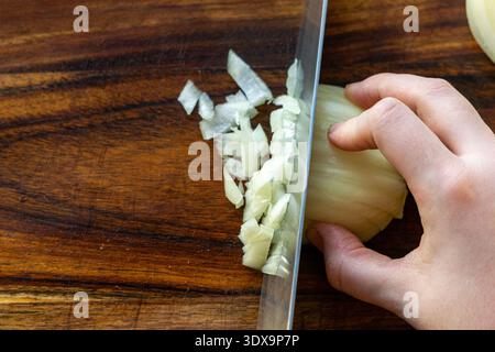 Gros plan d'une main hachant l'oignon blanc avec un couteau de cuisine sur une planche à découper en bois. Banque D'Images