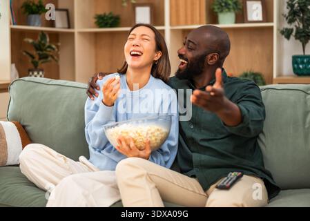 Un joyeux couple multiracial est assis étroitement ensemble sur un canapé vert, riant et embrassant tout en regardant la télévision et en mangeant du pop-corn. Banque D'Images
