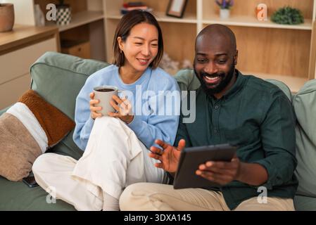 Couple interracial heureux partage un moment joyeux tout en se relaxant à la maison. Ils boivent du café, parlent et utilisent une tablette numérique. Banque D'Images