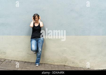 Femme blonde joyeuse appréciant la musique avec des écouteurs contre le mur de la ville Banque D'Images