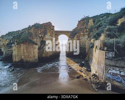 Vue aérienne des falaises escarpées embrassent une plage cachée, reliée par un ancien pont voûtant sur la mer turquoise, Lagos, district de Faro, Portugal. Banque D'Images