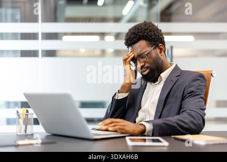 Jeune homme éprouvant de la frustration au travail et de l'épuisement mental tout en travaillant sur un ordinateur portable, représentant le stress, l'épuisement professionnel et la pression dans un environnement d'entreprise Banque D'Images