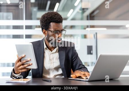 Jeune homme d'affaires en lunettes et costume concentré sur le multitâche à un bureau moderne, tapant sur un ordinateur portable tout en tenant une tablette numérique, la planification de la stratégie et l'analyse des données Banque D'Images