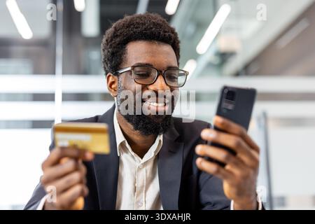 Homme souriant trouvant du plaisir dans l'achat en ligne, tenant une carte de crédit et un téléphone portable pour un paiement numérique sécurisé, représentant le e-commerce moderne Banque D'Images