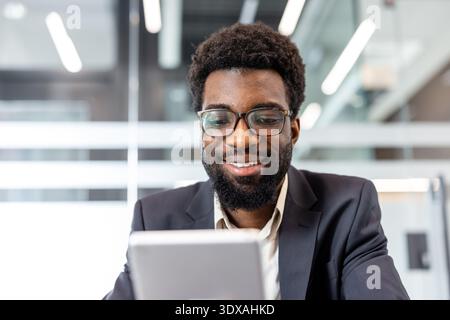 Jeune homme portant des lunettes et un costume de travail en toute confiance travaillant et souriant tout en utilisant une tablette numérique pour la communication professionnelle dans un espace de travail contemporain lumineux Banque D'Images