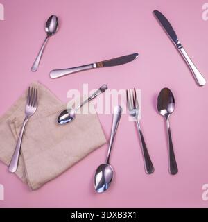 Une collection de couverts en acier inoxydable disposés sur un fond rose. Comprend des fourchettes, des cuillères et un couteau, avec une serviette beige en dessous. Banque D'Images
