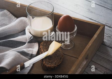 Un petit déjeuner basique : oeuf dur dans un coquetier avec une tranche de pain avec du beurre et un verre de lait sur un plateau en bois gros plan Banque D'Images