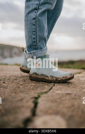 Une personne portant des jeans bleus et des bottes marche sur une plage rocheuse. Les bottes sont sales et boueuses, et les pieds de la personne sont couverts de boue. La scène Banque D'Images