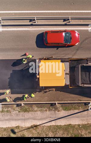 Travaux routiers : gros plan du finisseur d'asphalte et d'un tombereau avec de l'asphalte frais chaud en cours de déchargement et des ouvriers posant de l'asphalte frais sur l'autoroute A. Banque D'Images