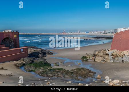 Vue panoramique sur la plage de Matosinhos au Portugal avec des gens qui se promènent sur le sable, l'horizon de la ville et le port de Leixões sous un ciel bleu clair. Banque D'Images