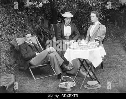 Photographie victorienne d'une scène de thé en plein air formelle de la fin de l'époque victorienne ou du début de l'ère édouardienne, mettant en scène un homme et deux femmes vêtus d'une tenue d'époque fidèle assis dans un jardin. Banque D'Images