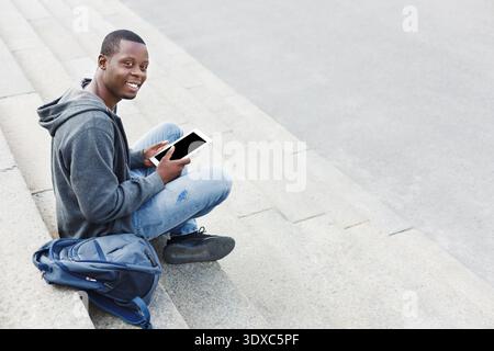 Jeune homme est assis sur les marches avec tablette et sac à dos tout en souriant Banque D'Images