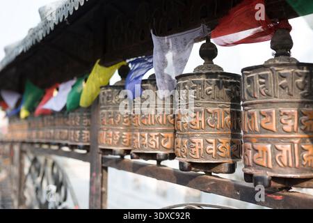 Rangée de roues à prières tibétaines en bronze avec drapeaux flottant au Stupa Swayambhunath, Katmandou, Népal. Banque D'Images