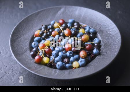 Baies d'été mélangées dans un bol gris, composition de vue de dessus sur une table sombre. Myrtilles, mûres et groseilles à maquereau, récolte biologique Banque D'Images