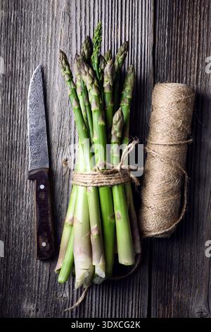 Bouquet de lances d'asperges vertes avec couteau et ficelle de cuisine sur fond de bois foncé. Légumes crus frais dans un style alimentaire minimal. Plat, vue de dessus Banque D'Images