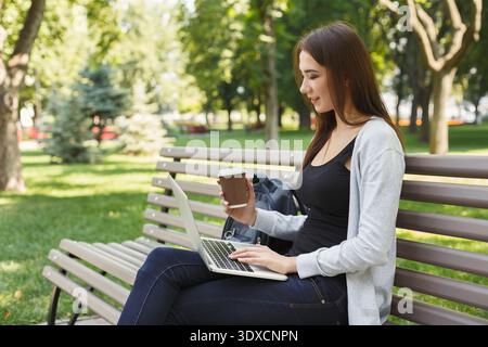 Jeune femme travaille sur ordinateur portable tout en étant assise sur un banc dans un parc avec du café dans sa main Banque D'Images