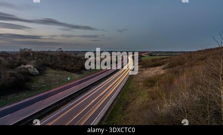 Vue longue exposition d'une autoroute courbe au crépuscule avec des traînées légères de passage de voitures à travers un paysage rural sous un ciel bleu nuageux, vu d'un el Banque D'Images