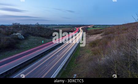 Vue crépusculaire longue exposition d'une autoroute incurvée avec des sentiers de lumière rouge et blanche à travers le paysage rural, vu d'un passage supérieur, avec ciel ouvert et policier Banque D'Images